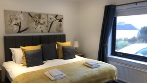 A neatly made bed with a yellow blanket, a headboard with three flower paintings, a bedside lamp, and a window with a view of a car and landscape. - Glendaruel Bed & Breakfast (Portree)
