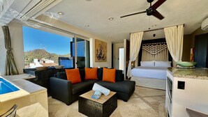 A hotel room with a balcony, a bed, a sofa with orange cushions, a dining area, and a ceiling fan.