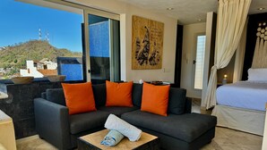 A modern living room with a dark grey sofa, orange cushions, and a wooden coffee table. A large window offers a view of a cityscape and a hill.