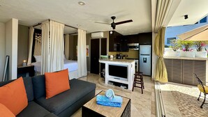A modern living room with a sofa, a coffee table, and a TV. There is a kitchen area with a refrigerator and a microwave. The room has a ceiling fan and curtains.