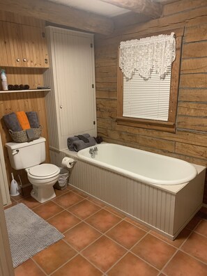 Un baño con inodoro y bañera blancos, una ventana con cortina de encaje blanco y paredes de madera.