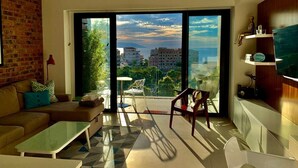 Een moderne woonkamer met een bank, salontafel en stoelen, een grote schuifglazen deur die uitkomt op een balkon met uitzicht over de stad.