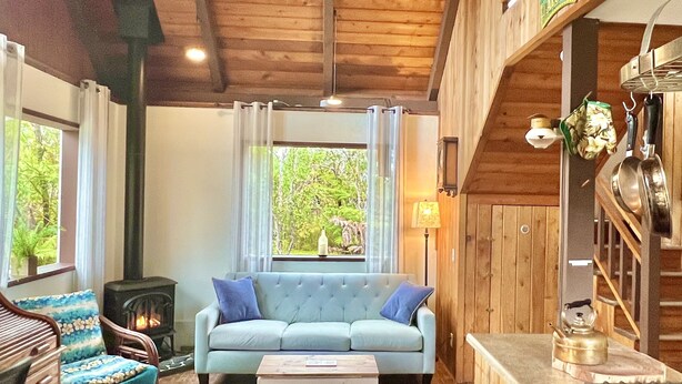 Un salon chaleureux avec un canapé bleu clair, une cheminée, une table à manger et un coin cuisine doté de meubles en bois et d’une cuisinière à gaz.