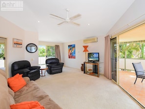 A living room with a sofa, two black leather chairs, a TV on a glass table, a ceiling fan, and a balcony with a view of palm trees. - Fabulous Pet Friendly Family Home - 3 Carribean Court (Castaways Beach)
