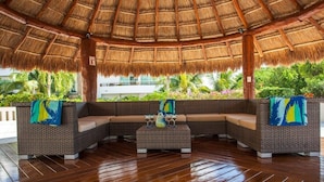 An outdoor seating area with a thatched roof, wicker furniture, and a view of lush greenery. - Great 1BR Condo w/ Pool, Near Beach! AC! (Playa Del Carmen)