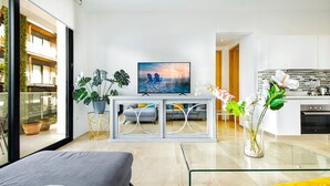 Un salon moderne avec un canapé, une table basse en verre et un téléviseur posé sur un meuble blanc. On y trouve également un coin cuisine avec une cuisinière et des placards.