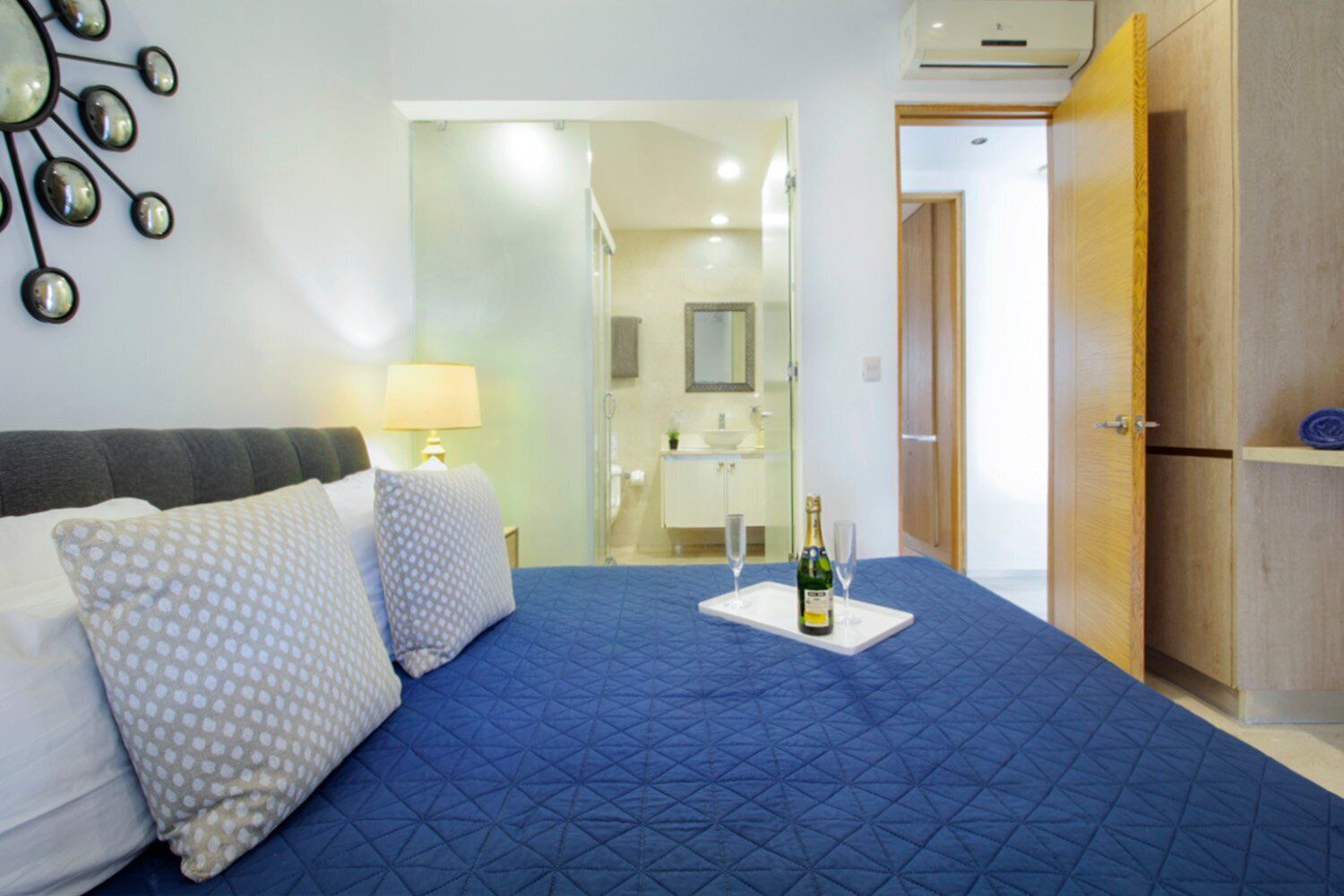 Un dormitorio con una cama grande, una mesita de noche con lámpara, una botella de champán y un espejo en la pared.