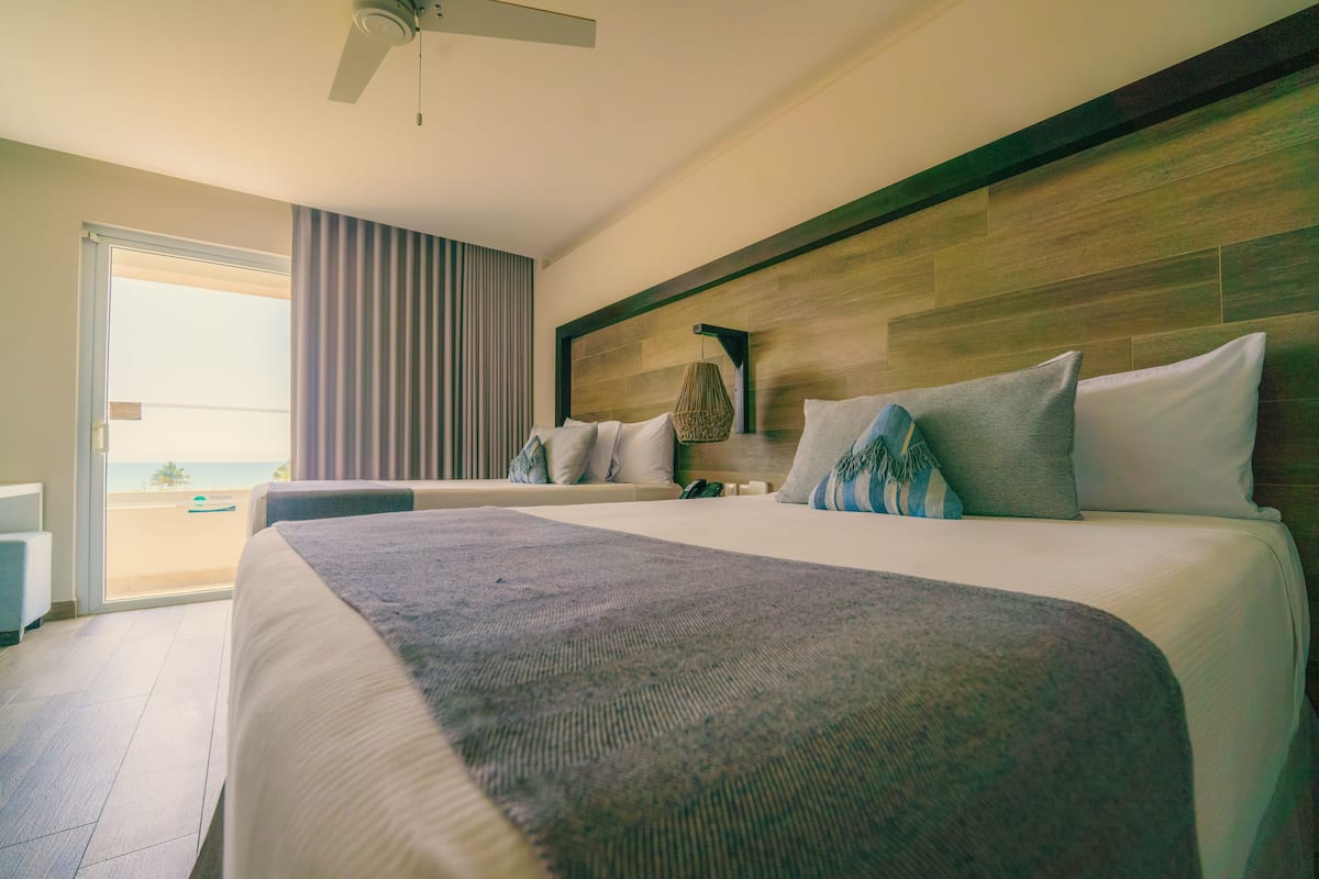 A hotel room with a large bed, a ceiling fan, and a view of the beach through the sliding glass door.