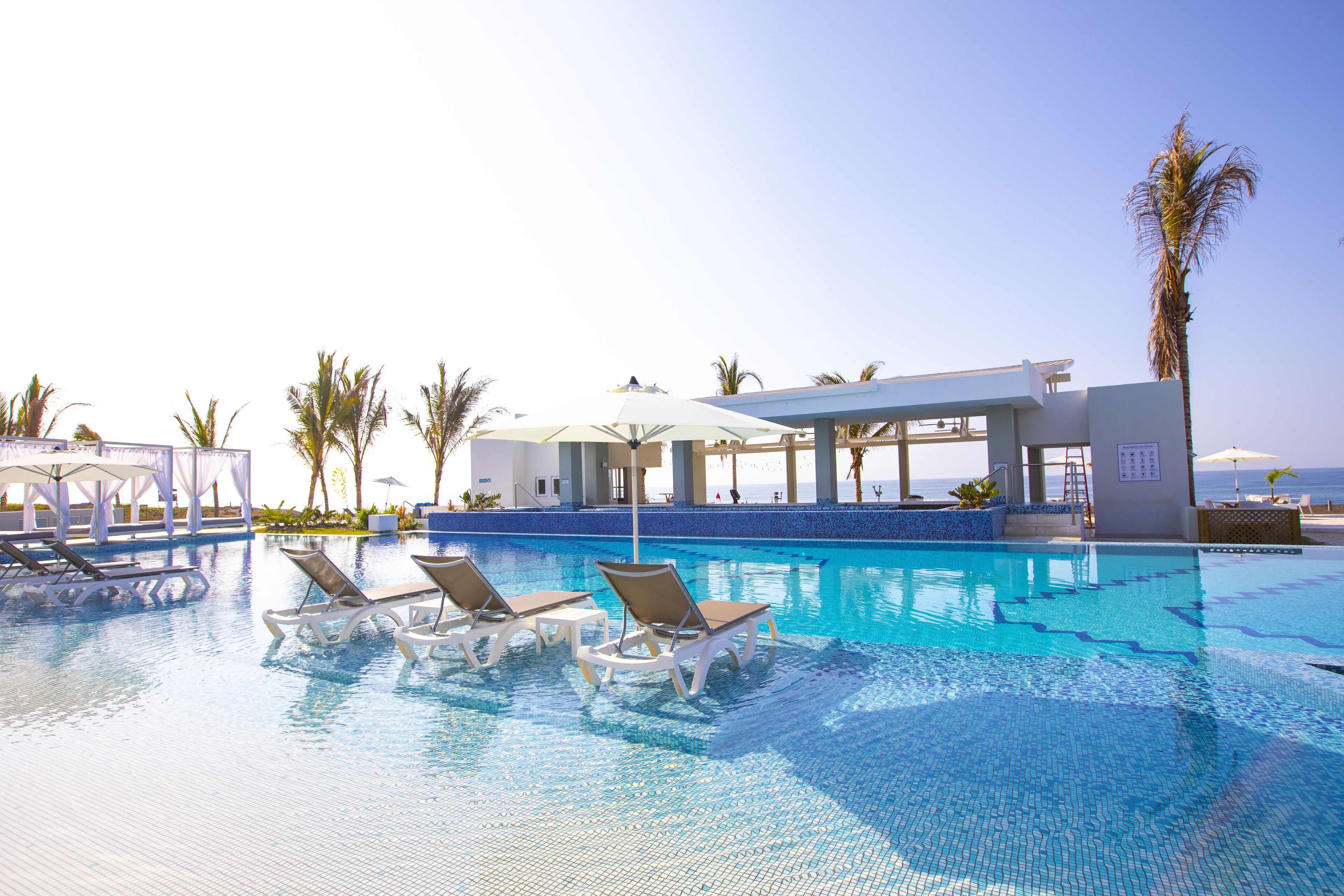 Zona de piscina con sillones reclinables, sombrillas y un edificio con vista al océano.
