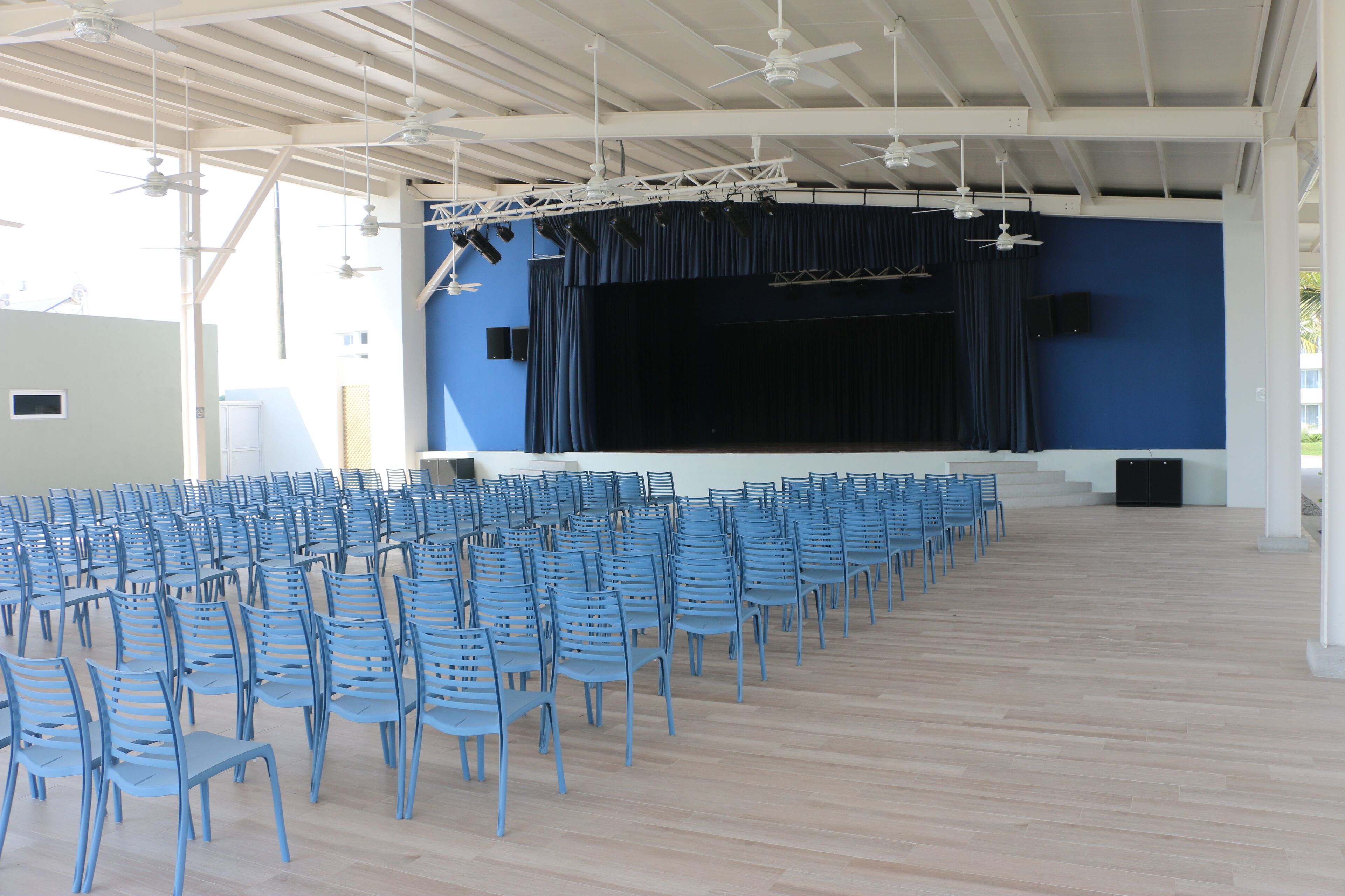 Un espacio interior con un escenario, filas de sillas azules, ventiladores de techo y un piso de color claro.