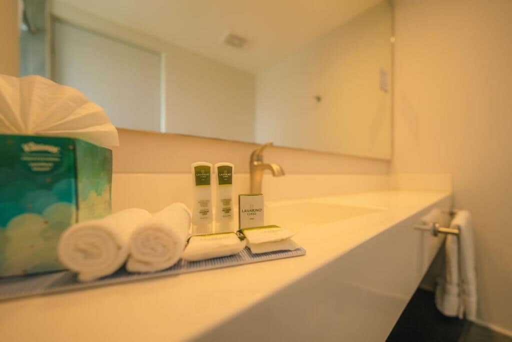 A bathroom with a white sink, a mirror, and toiletries including a green box, two bottles, and rolled towels.