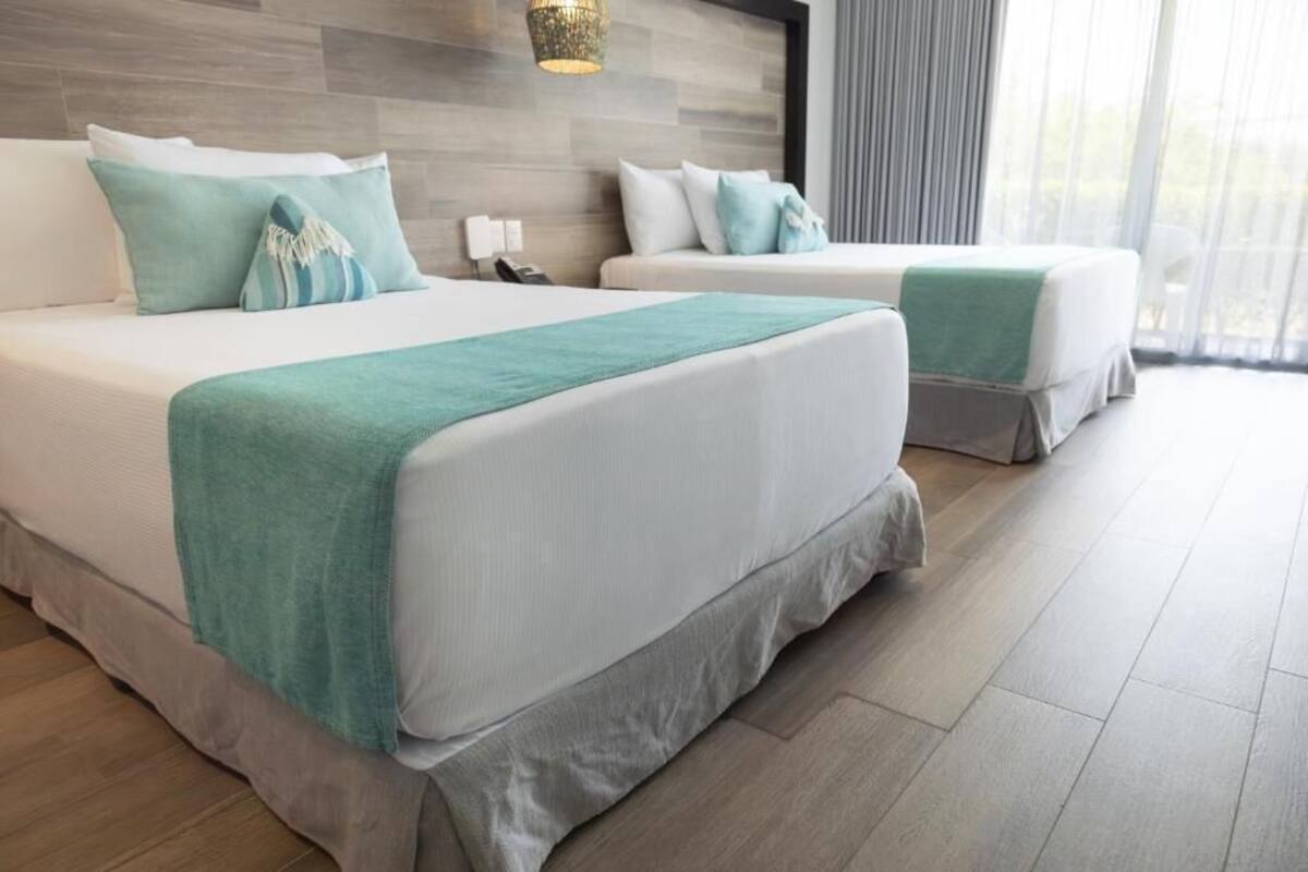 A hotel room with two beds, a wooden headboard, a bedside table with a lamp, and a large window with curtains.