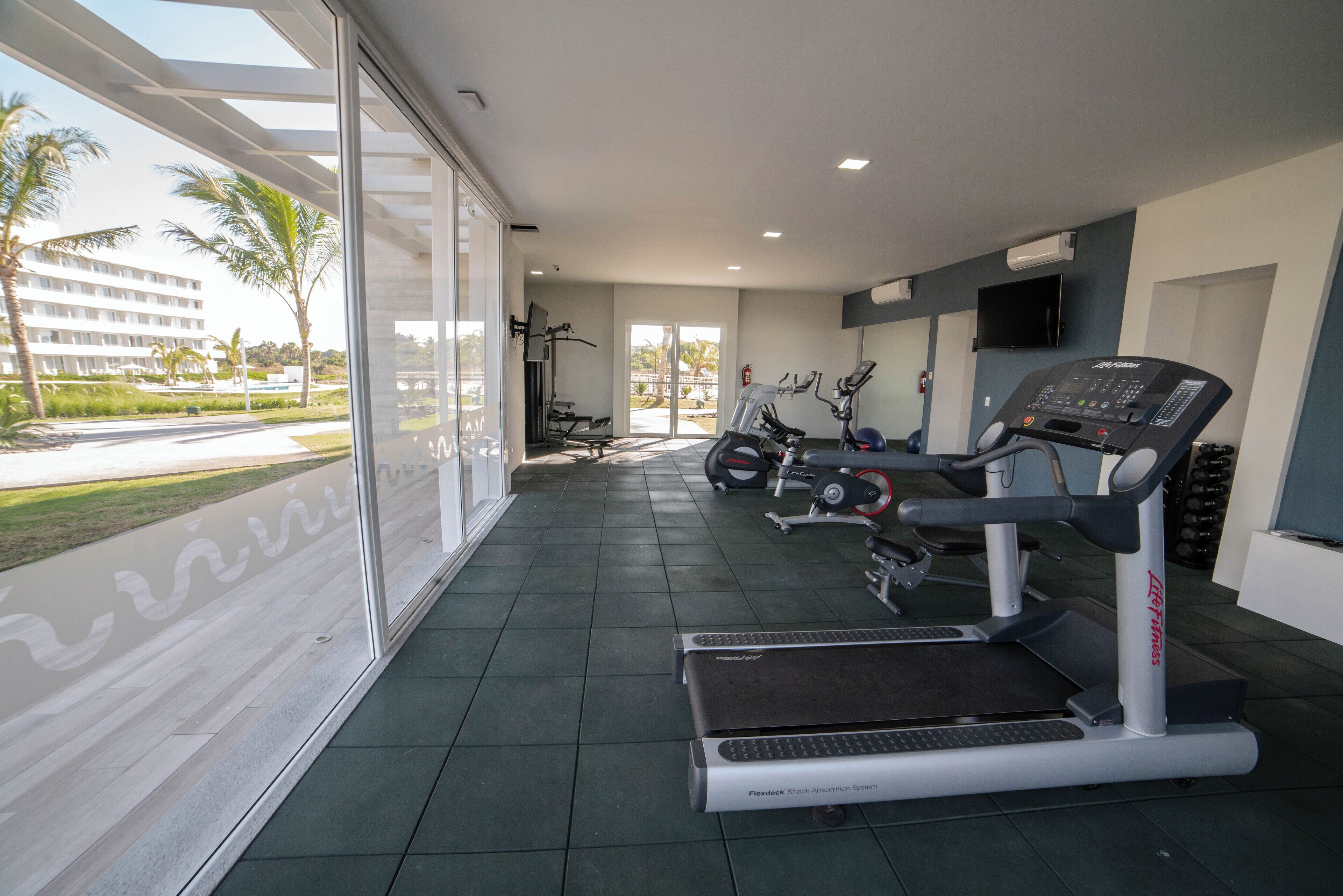 Ein modernes Fitnessstudio mit Laufbändern, Krafttrainingsgeräten und einem großen Fenster mit Blick auf Palmen und ein Gebäude.