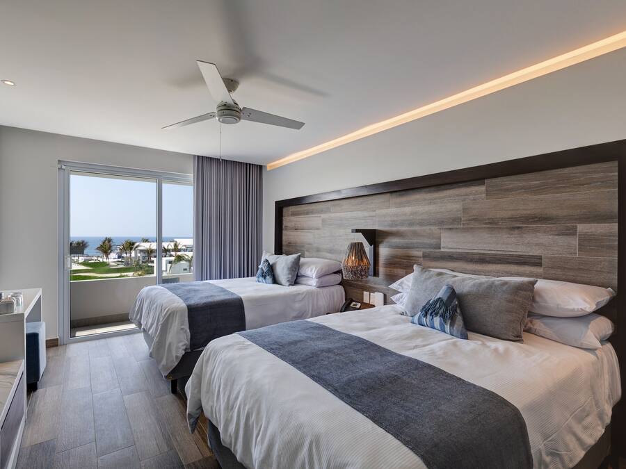 Una habitación de hotel moderna con dos camas, un ventilador de techo, un ventanal con vistas y una mesita de noche con lámpara.