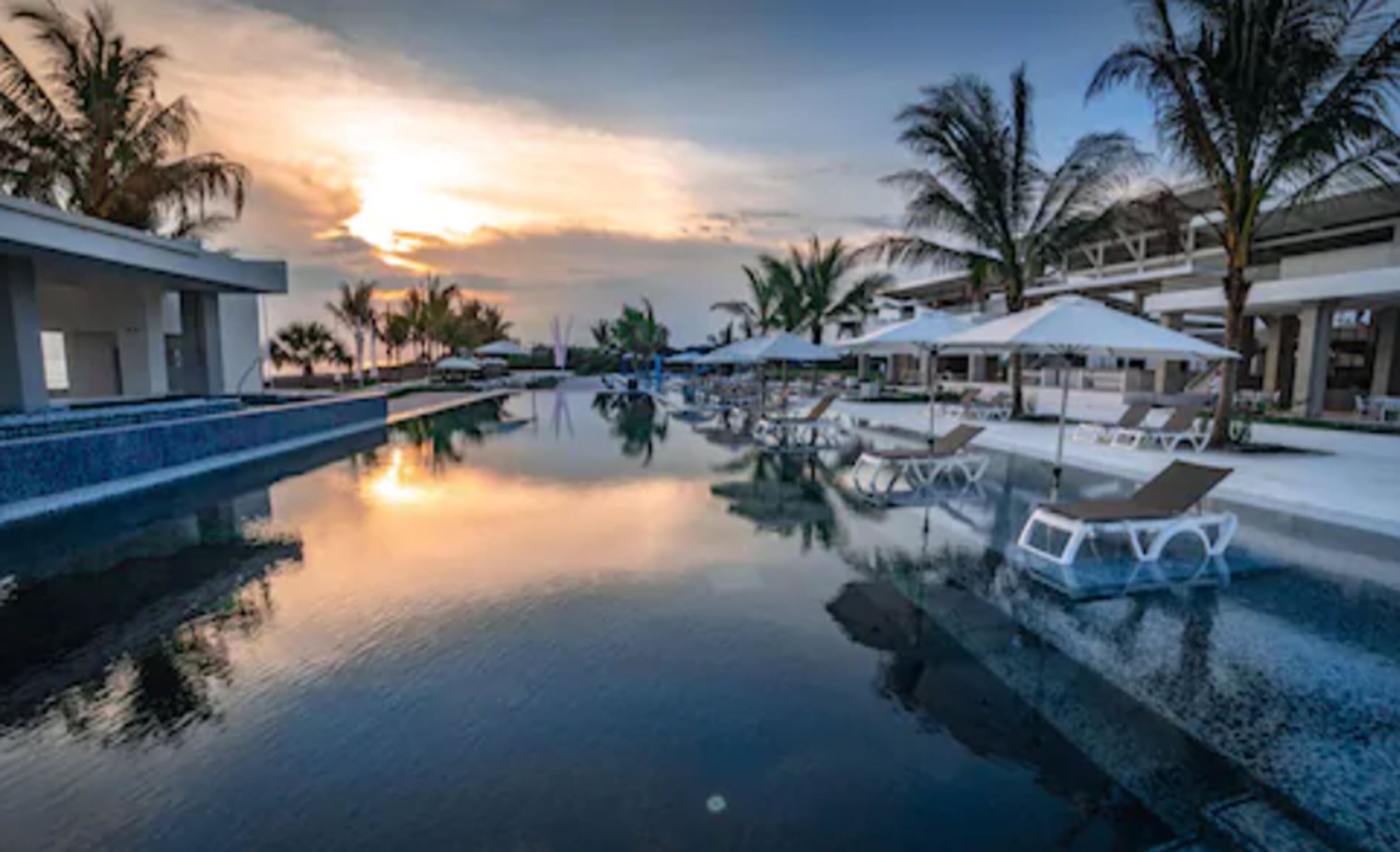 A serene pool area with lounge chairs and umbrellas, surrounded by palm trees and modern buildings.