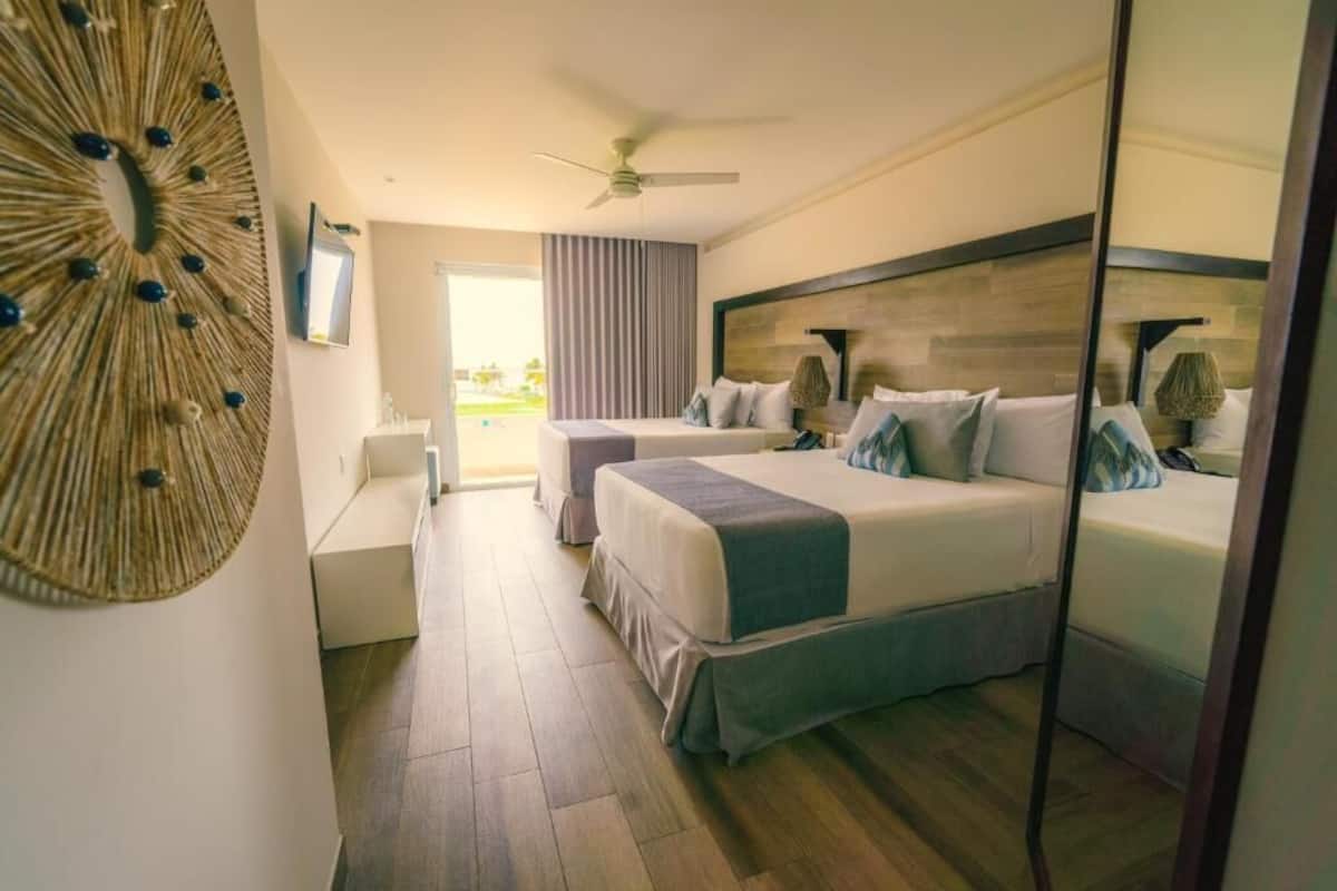 A hotel room with two beds, a ceiling fan, a large wall decoration, and a view of the outdoors.