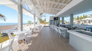 Un área de bar al aire libre con sillas y mesas blancas, una terraza de madera y vistas a la playa.