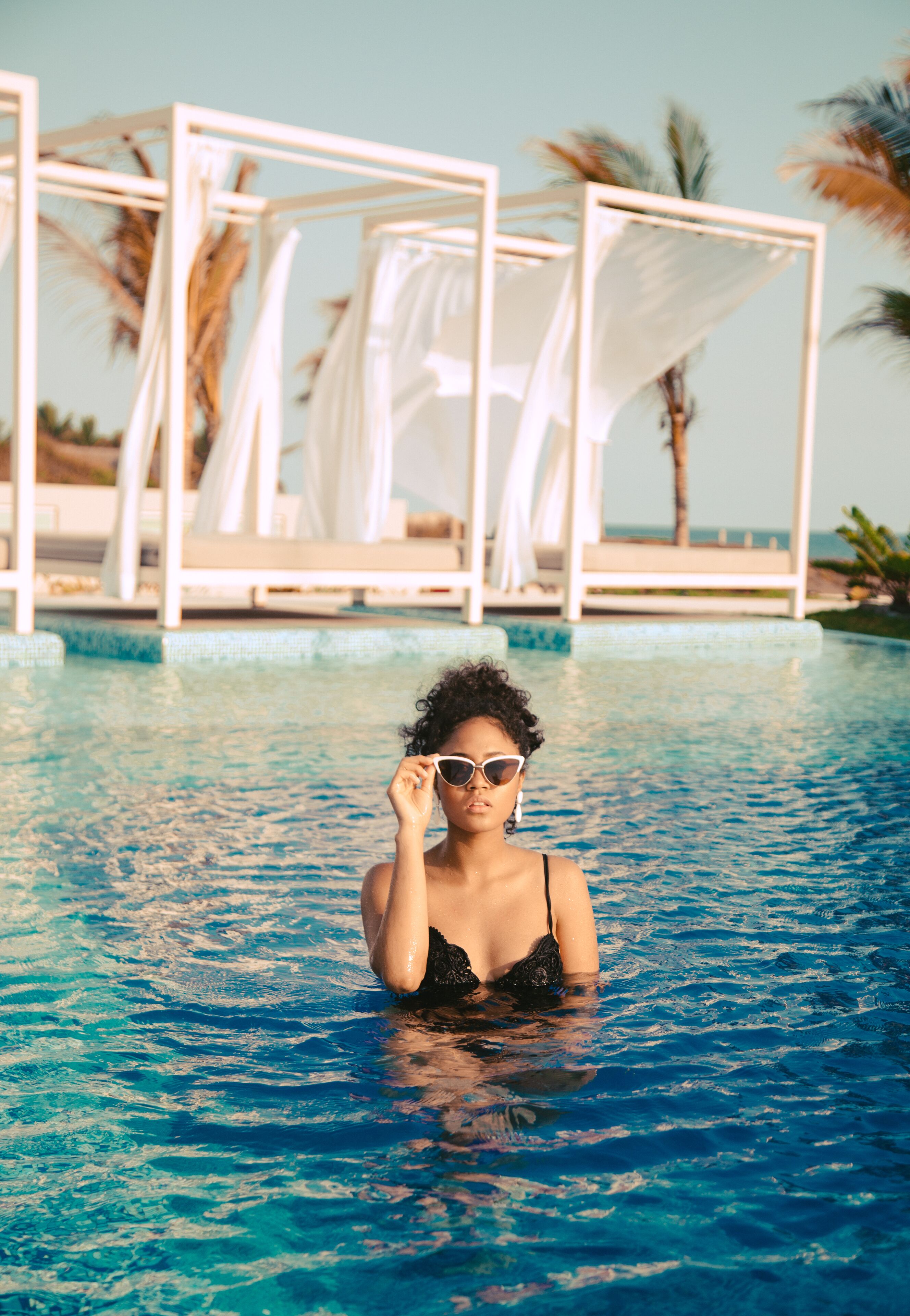 A pool area with a person in the water, lounge chairs, and palm trees in the background.