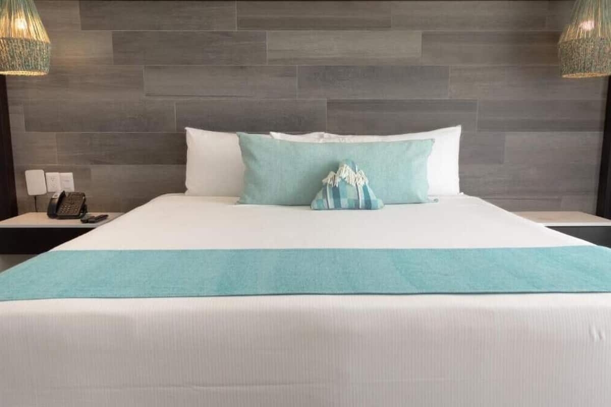 A hotel room with a large bed, a teal throw blanket, and a checkered pillow. There are two bedside tables with lamps and a phone on one of them.