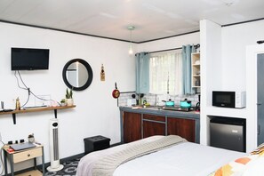 A compact bedroom with a bed, bedside table, and a small desk with a chair. A TV is mounted on the wall, and a window with curtains is visible.