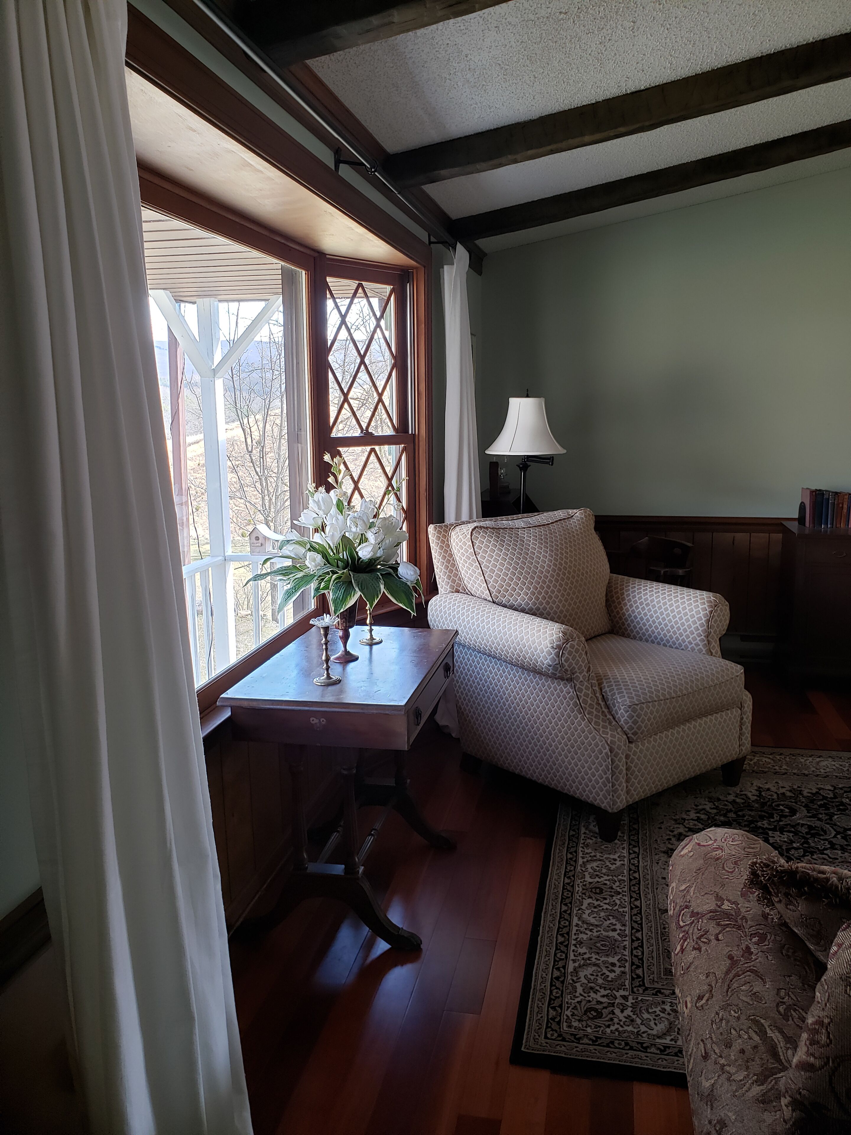Una habitación acogedora con una mesa de madera, un sillón estampado, una lámpara y vistas a través de una ventana con cortinas.