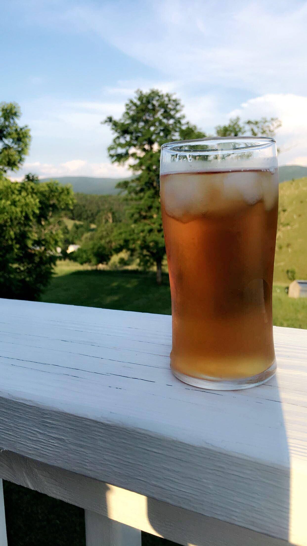A glass of iced tea on a white wooden deck with a view of trees and hills in the background.