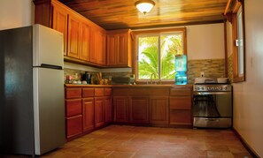 A kitchen with wooden cabinets, a refrigerator, a stove, and a window with a view of palm trees. - Chakra Le Esconddida House (San Pedro)