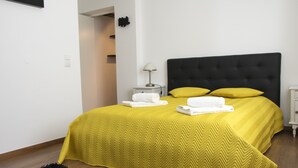 Un lit bien fait, recouvert d’une couvre-lit jaune, avec une tête de lit noire et deux oreillers. On y trouve une table de chevet blanche, sur laquelle se trouvent une lampe et une serviette blanche pliée. Les murs de la pièce sont blancs et un tapis noir recouvre le sol.