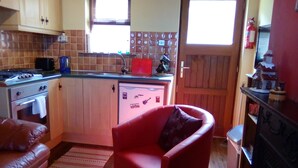 Cocina compacta con estufa, lavaplatos y refrigerador. Hay una silla roja en el centro y una alfombra pequeña en el suelo.