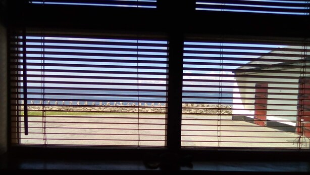 Una ventana con persianas horizontales, vista al océano y una pared de piedra.