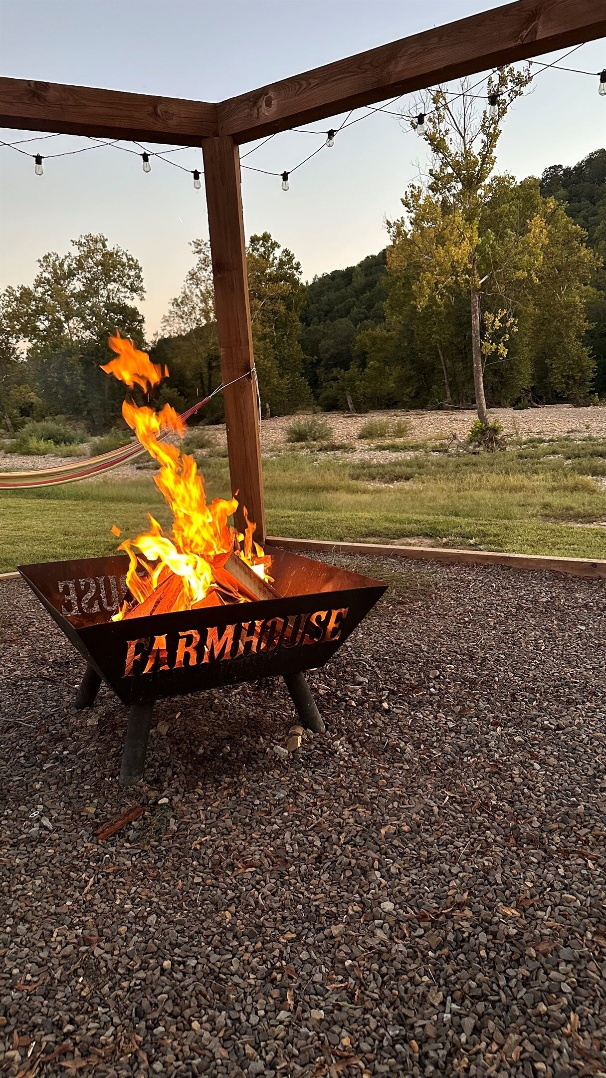 A metal fire pit with 'FARMHOUSE' engraved on it, surrounded by gravel and under a wooden pergola with string lights.
