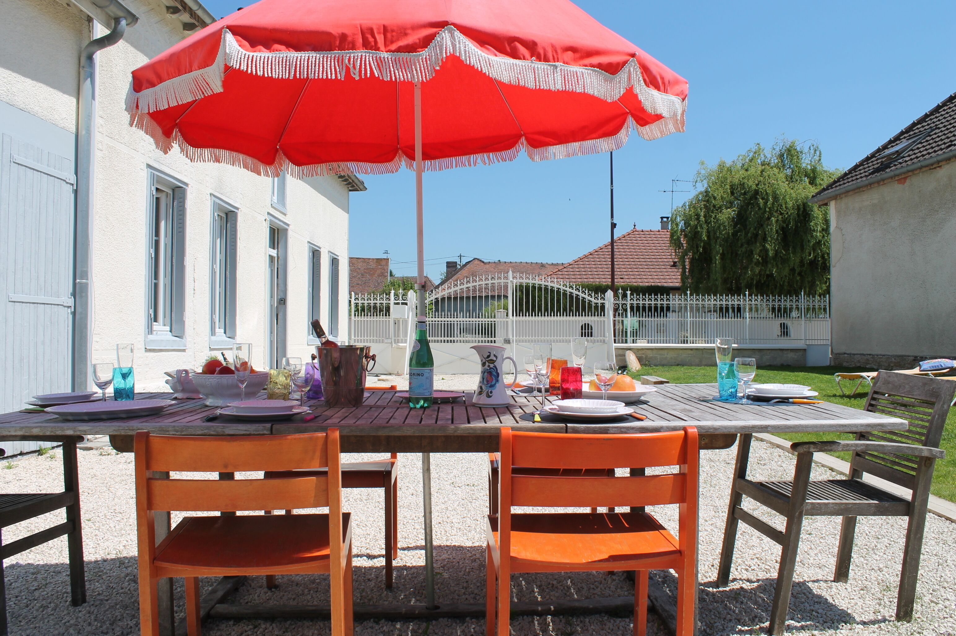 A table set outdoors with a red umbrella, surrounded by chairs, and various dining items.