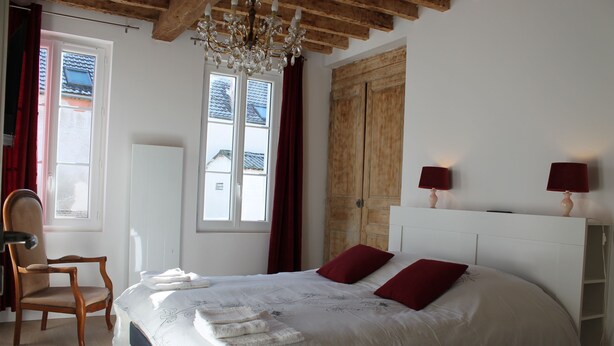 Ein Schlafzimmer mit einem großen Bett, zwei roten Kissen, einem Holzstuhl, einer Kristallleuchtre und einem Fenster mit Vorhängen.