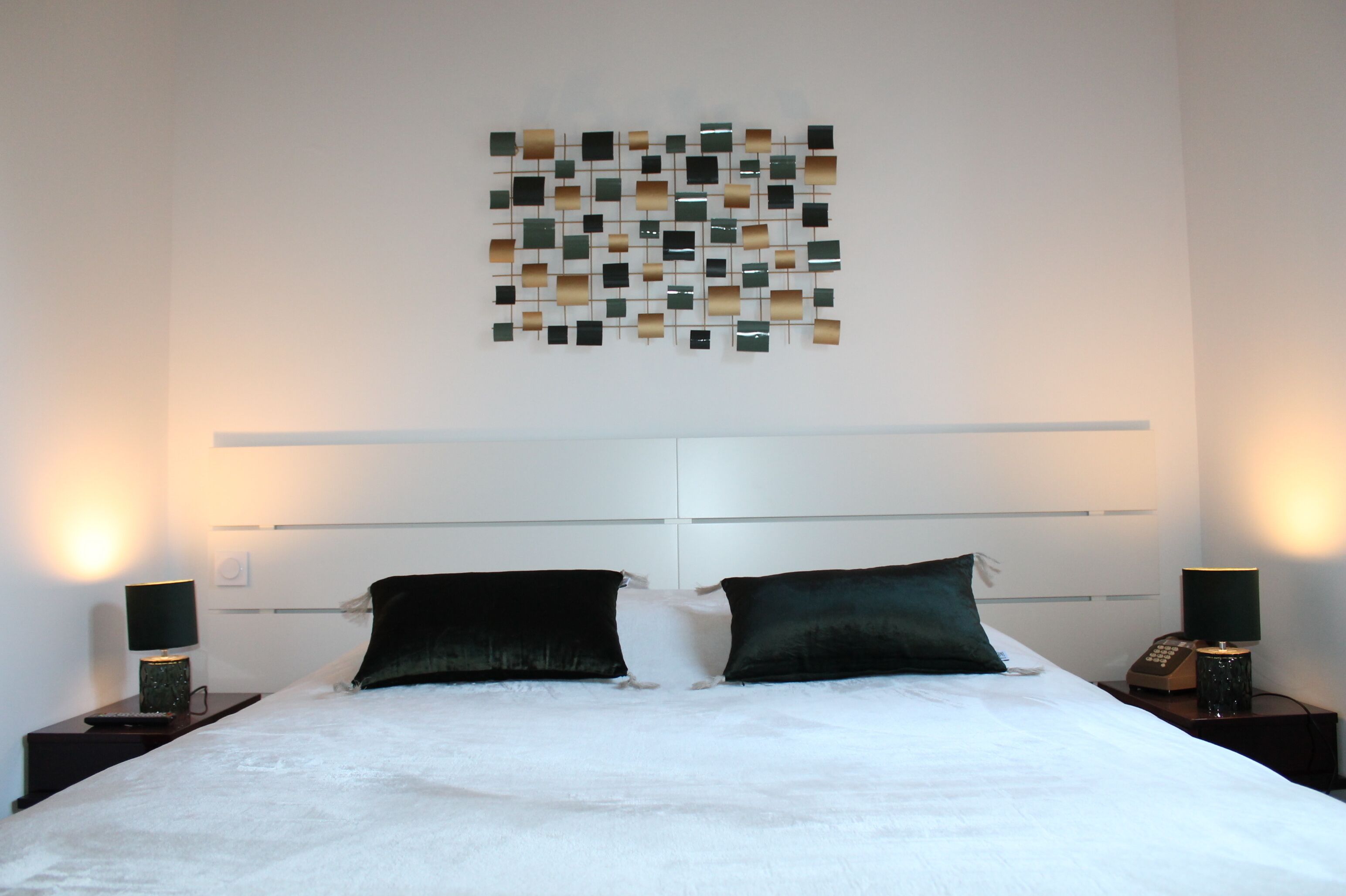 Una cama bien tendida con dos almohadas, una obra de arte en la pared encima y lámparas de noche a ambos lados.