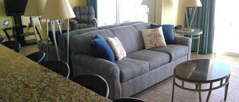 Una sala de estar con un sofá gris, dos sillones, una mesa de centro y un televisor. Hay dos lámparas, una silla y una mesa con una lámpara.