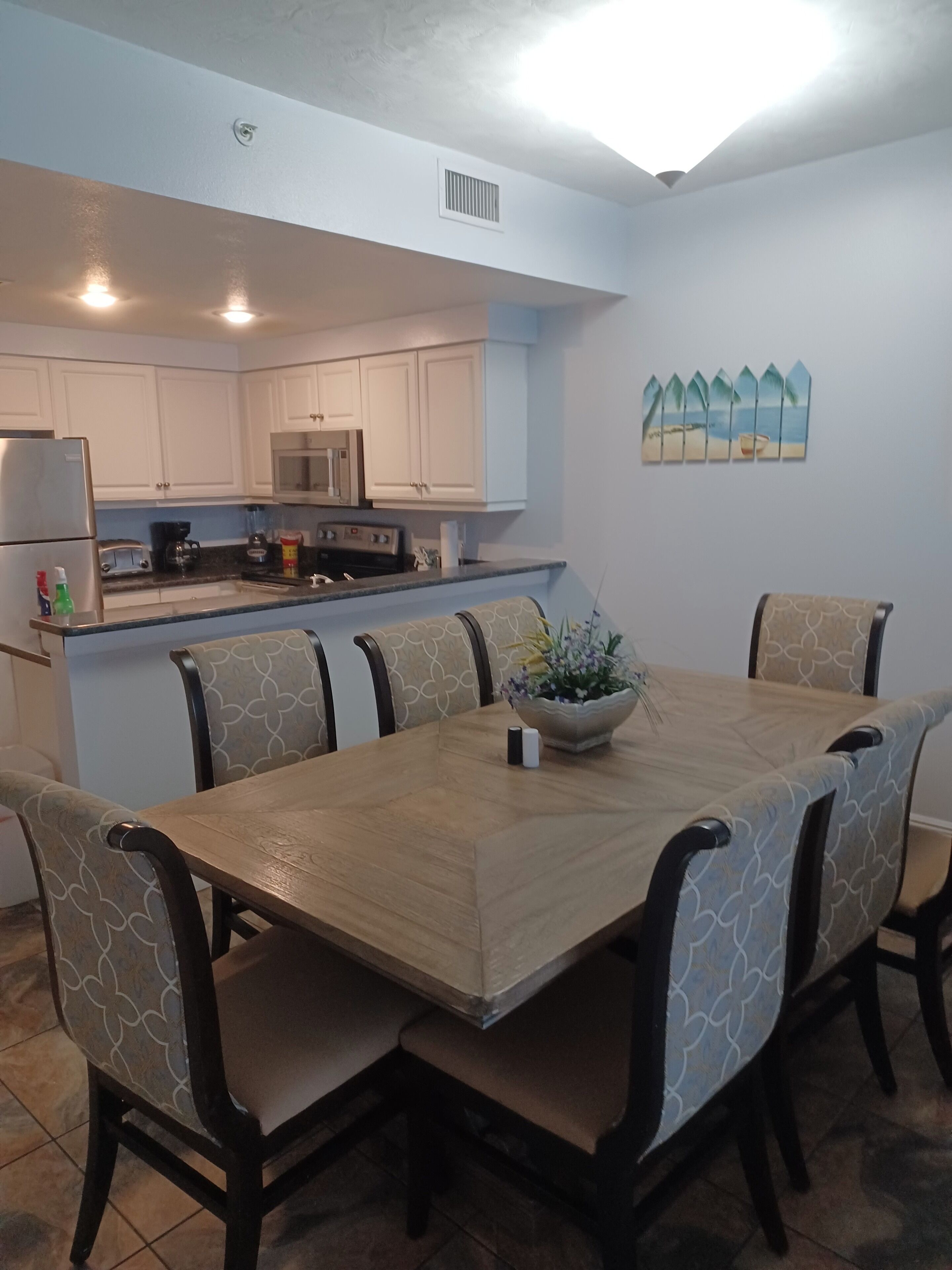 A dining area with a wooden table and patterned chairs, a kitchen with white cabinets, and a decorative wall piece.