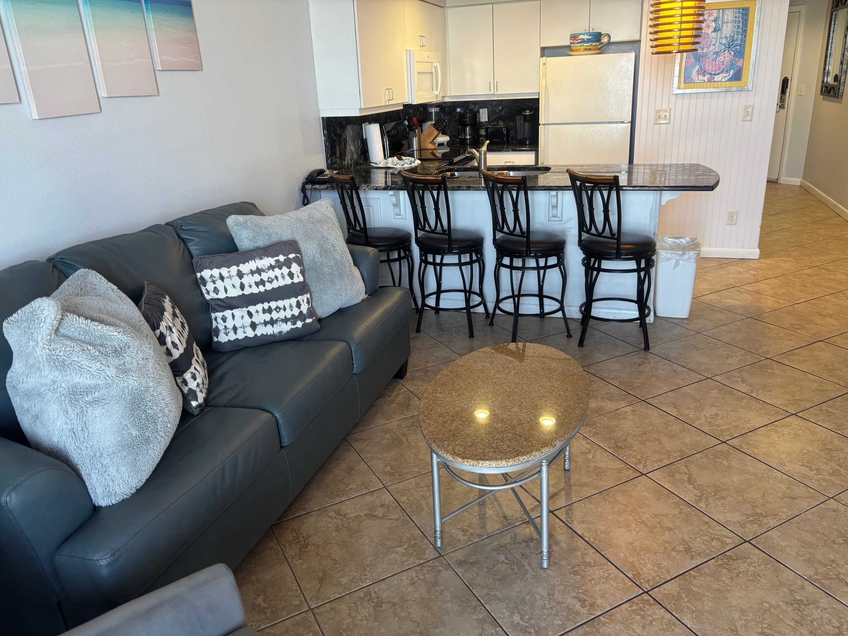 A modern living room with a black leather sofa, a round coffee table, and a kitchen area with a black countertop and white cabinets.