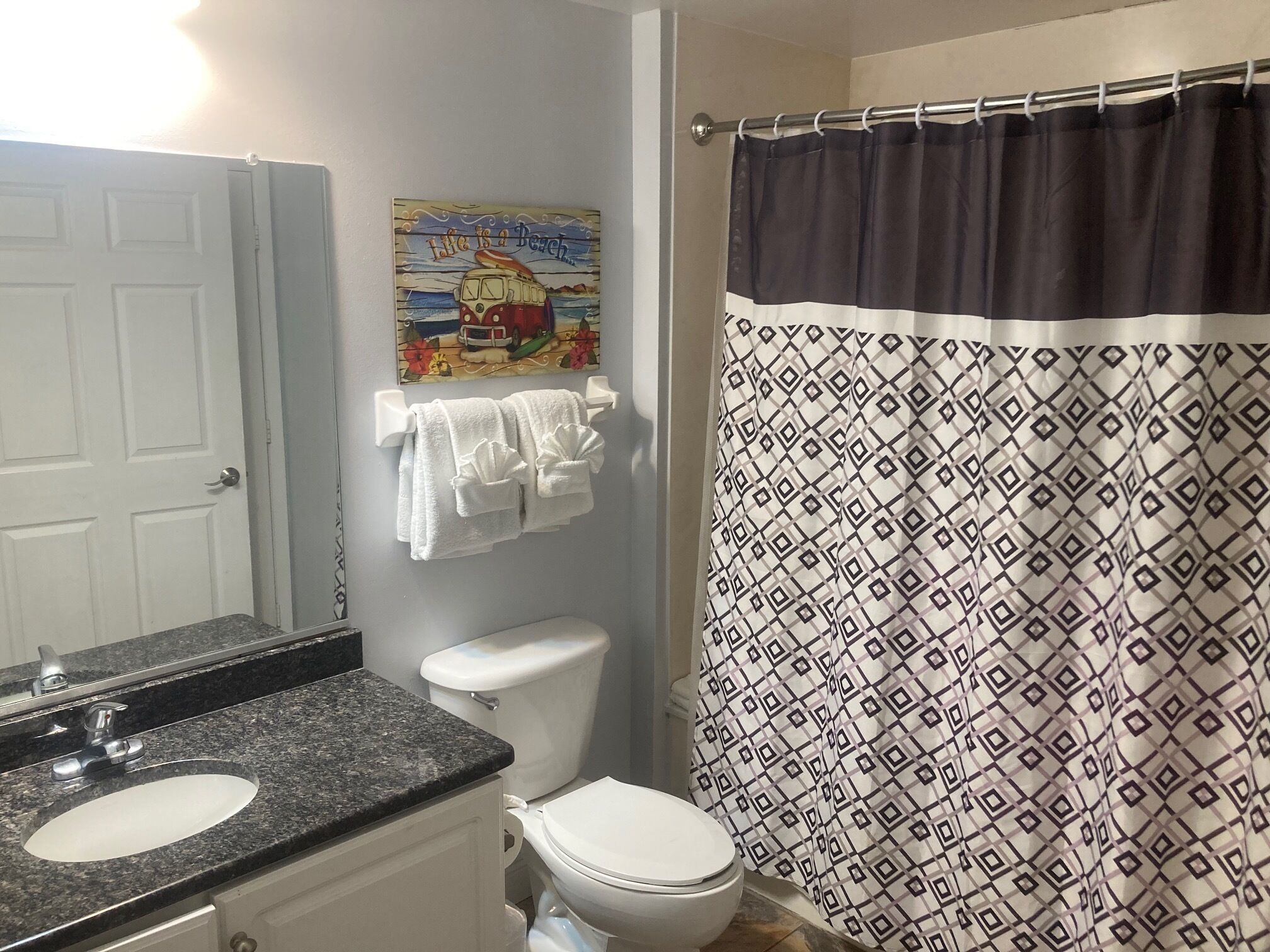 A bathroom with a white toilet, a sink with a granite countertop, a towel rack with white towels, a shower curtain with a geometric pattern, and a painting on the wall.