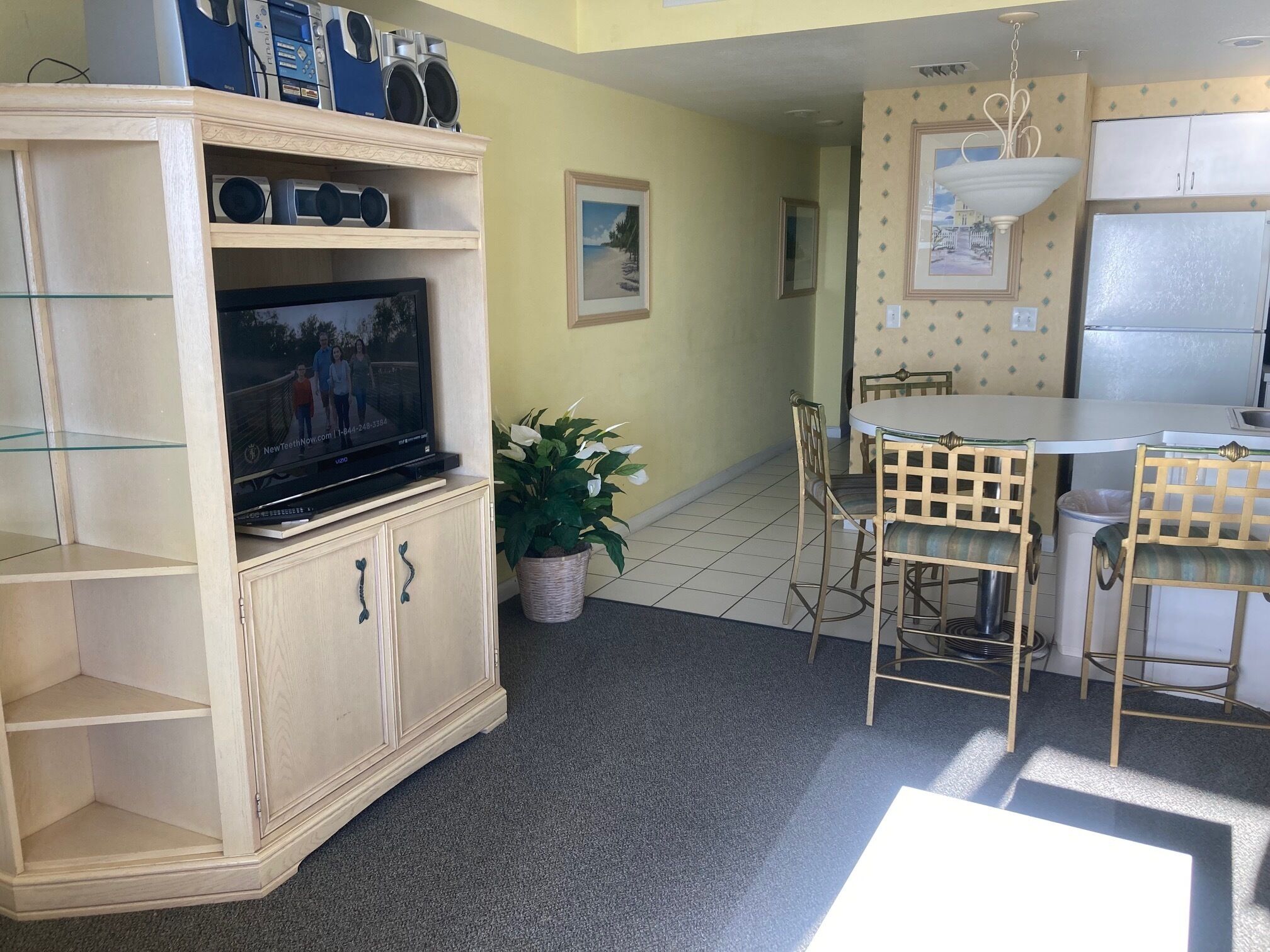 Un espacio habitable compacto con un televisor en un mueble de madera, una zona de comedor con mesa y sillas, y una cocina con refrigerador y alacenas.