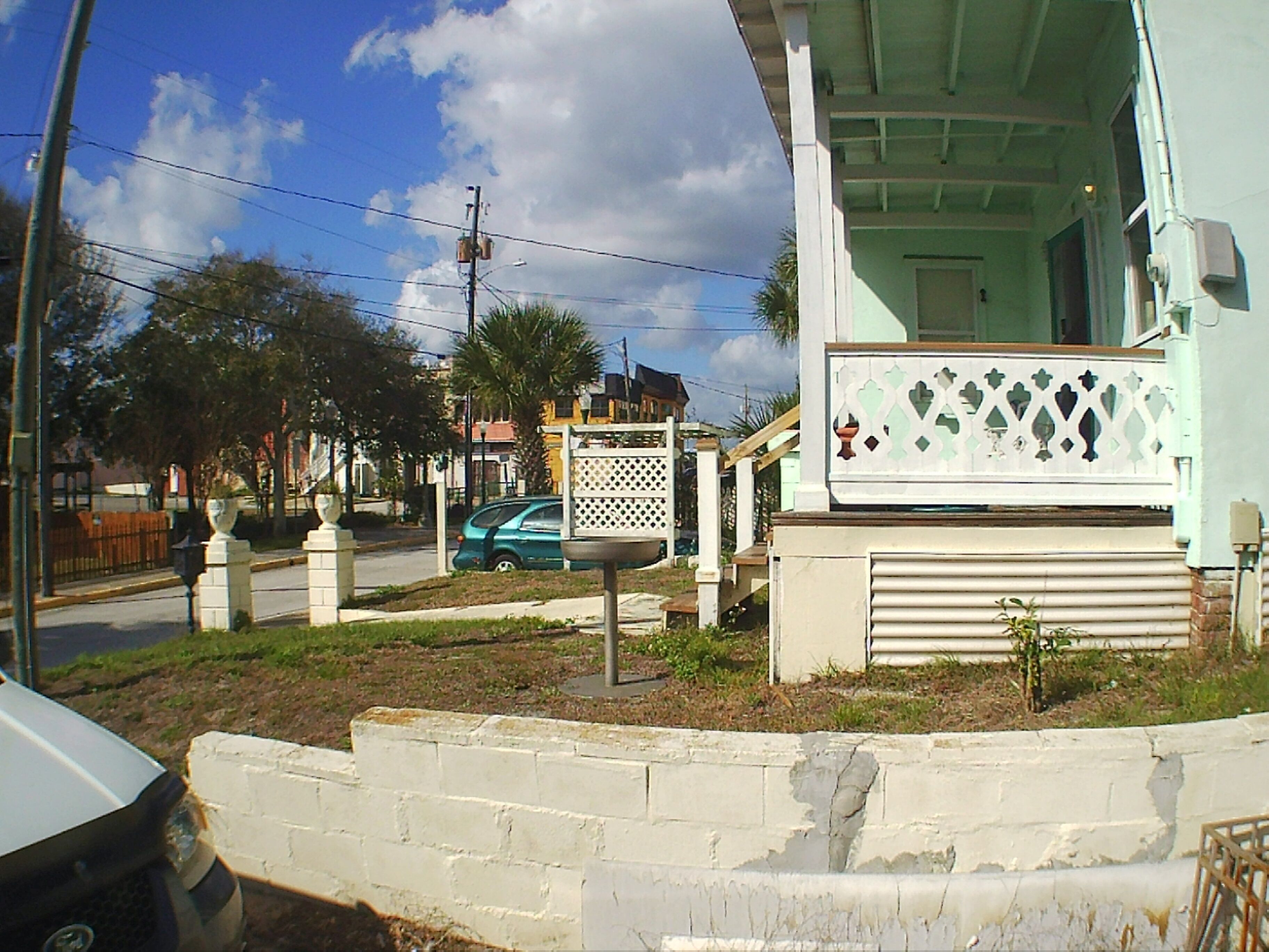 A residential area with a white house, a green car parked in front, and a street with trees and power lines.