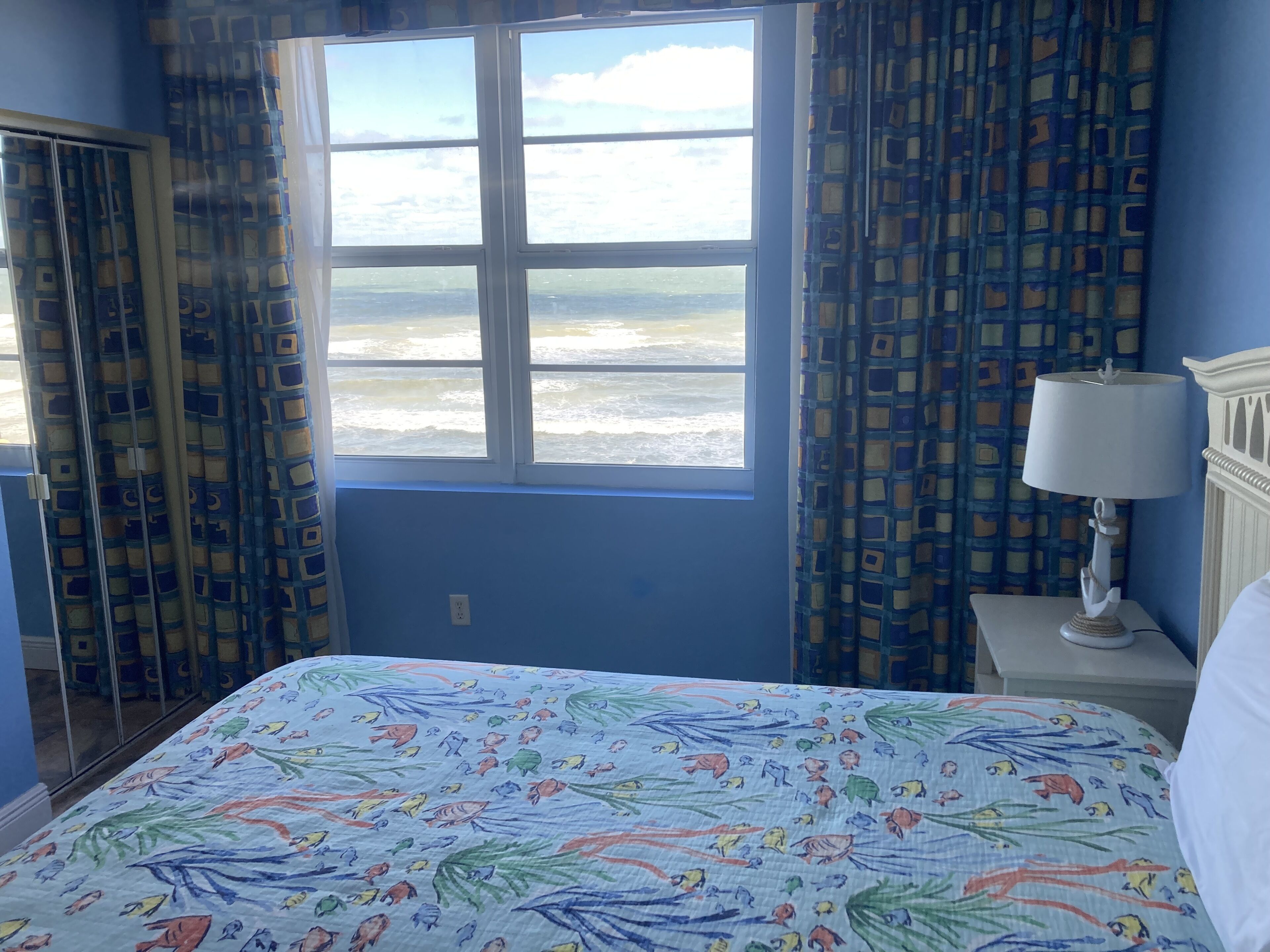 Quarto com cama, janela com vista para o mar, cortina estampada e mesa de cabeceira com luminária.