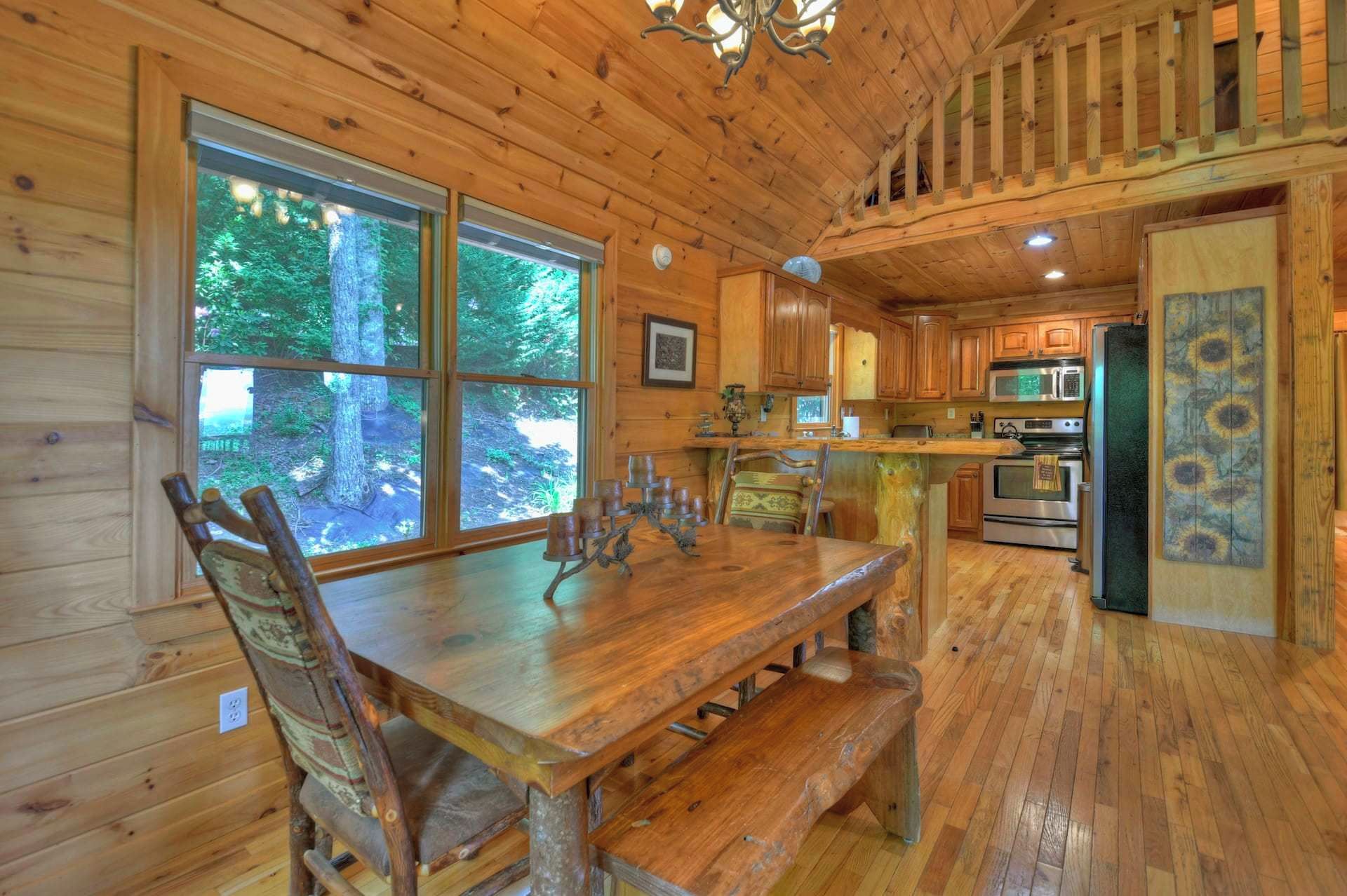A wooden cabin interior with a dining table, chairs, a kitchen area, and large windows overlooking a forest.
