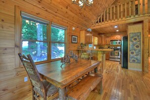 A wooden cabin interior with a dining table, chairs, a kitchen area, and large windows overlooking a forest.