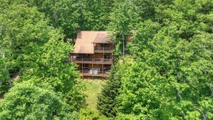 A two-story log cabin with a balcony, surrounded by dense forest. - Logging Memories | Mtn Views + Hot Tub + Games (Blue Ridge)