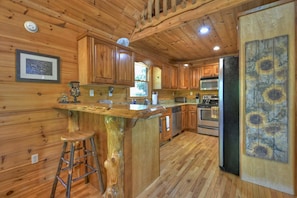 Interior de una cabaña de madera con cocina, que incluye armarios de madera, un taburete, un refrigerador y un cuadro de girasoles.