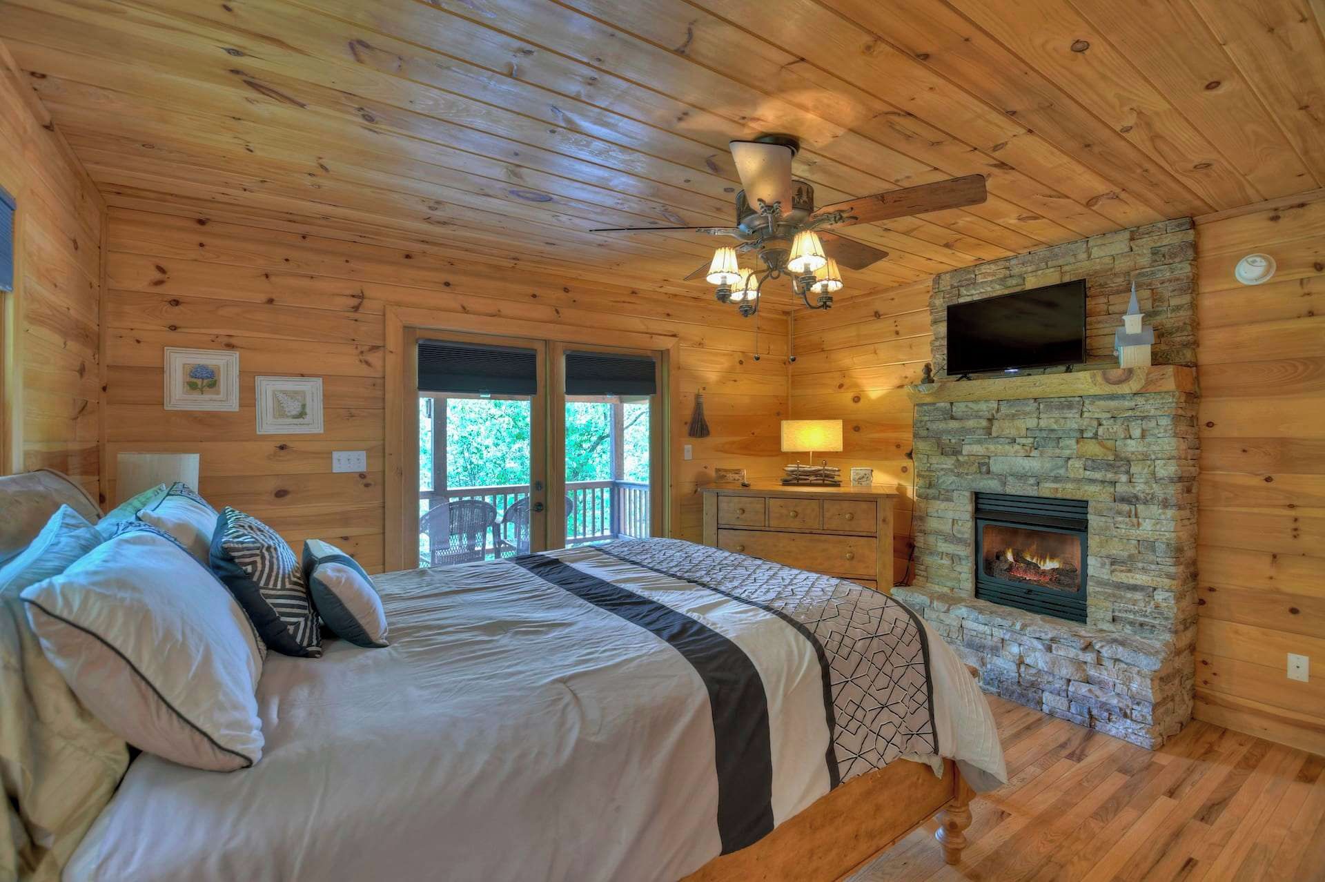 A wooden cabin bedroom with a bed, a stone fireplace, a television, and a ceiling fan.