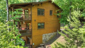 A two-story log cabin with a balcony, a stone wall, and a fire pit in the yard. - Logging Memories | Mtn Views + Hot Tub + Games (Blue Ridge)