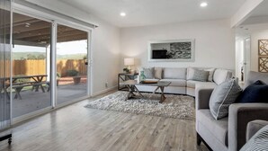 Ein modernes Wohnzimmer mit einer Schiebeglasdoor zur Terrasse, einem grauen Sofa, einem Couchtisch und einem an der Wand montierten Fernseher.