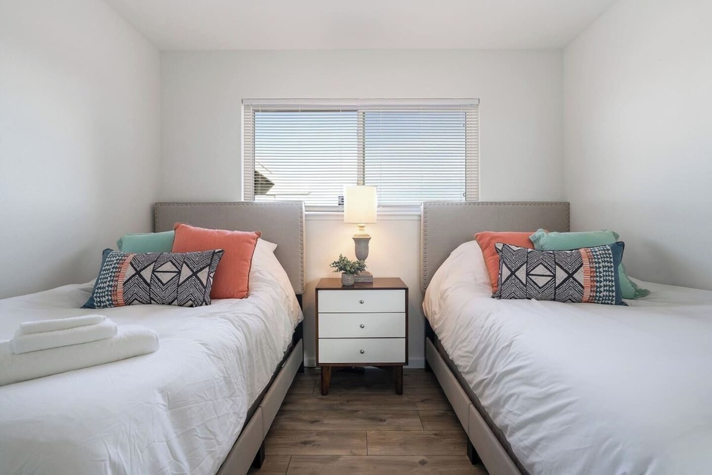 A twin bedroom with two beds, a nightstand with a lamp, and a window with blinds.