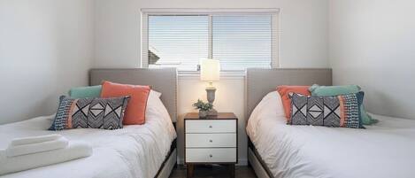 A twin bedroom with two beds, a nightstand with a lamp, and a window with blinds.